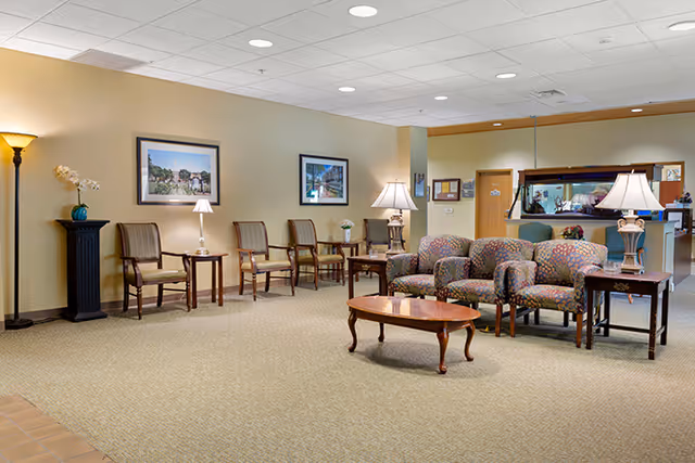 Spacious senior living lobby with upholstered chairs, wooden side tables and lamps, framed artwork, and a reception desk in the back.