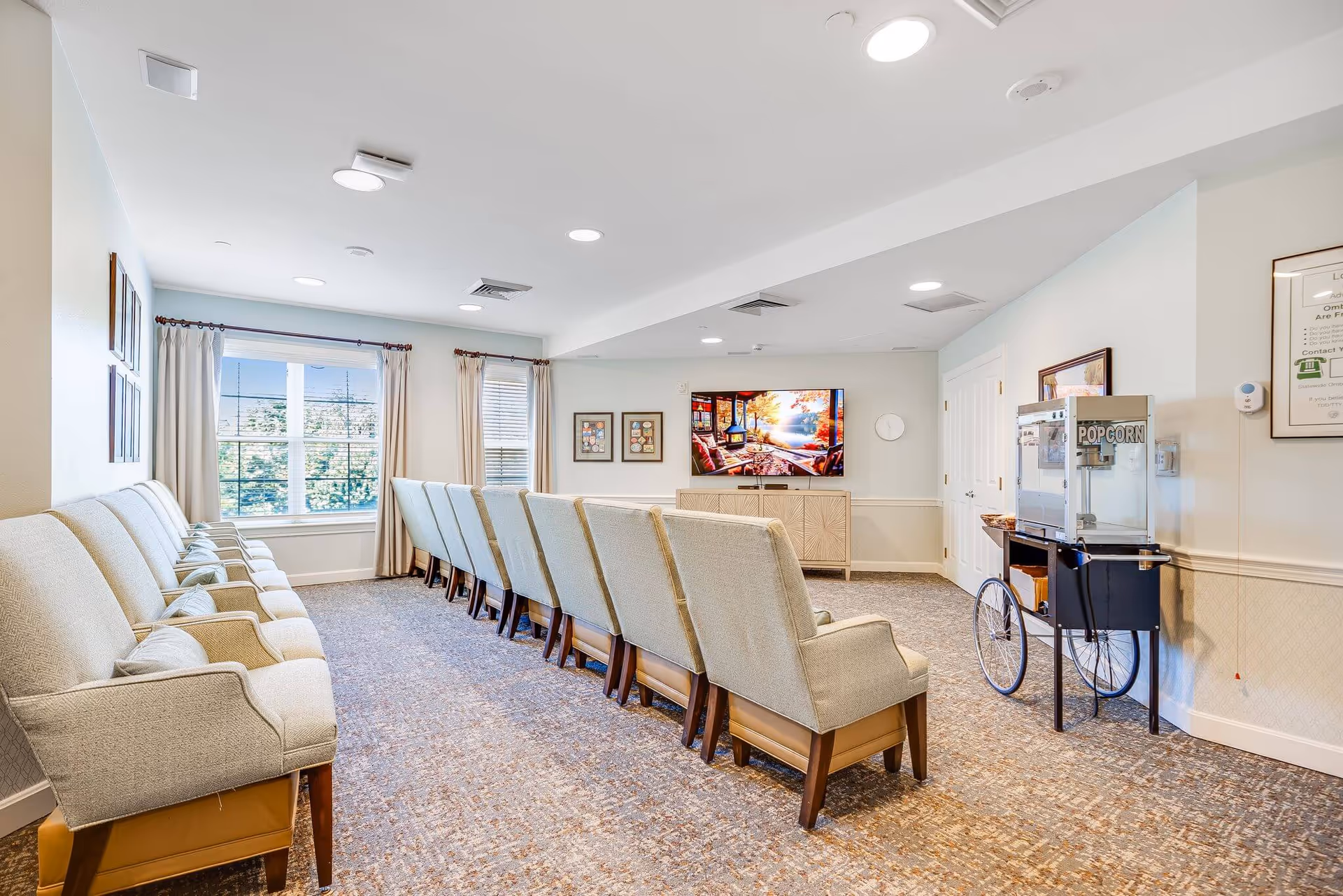 A bright and spacious common room with two rows of beige upholstered chairs facing a large flat-screen TV mounted on the wall. The room has large windows with curtains letting in natural light, a popcorn machine on a cart to the right, and framed artwork on the walls.