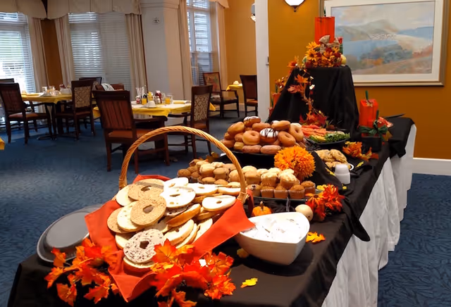 A buffet table set with a variety of breakfast pastries including bagels, muffins, and donuts, decorated with autumn-themed artificial leaves and flowers. In the background, there are dining tables with chairs, set with yellow tablecloths and condiments, in a well-lit room with large windows and beige curtains.