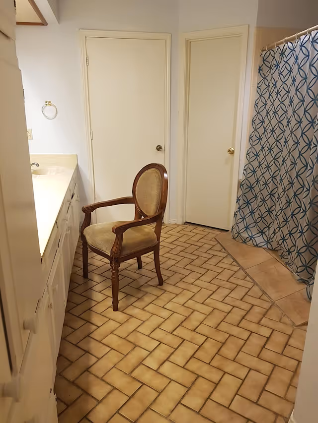 A bathroom with beige tile flooring arranged in a herringbone pattern, a wooden chair with cushioned seat and backrest placed near a long countertop with a sink. There are two closed white doors on the far wall, and a shower area with a blue and white patterned shower curtain on the right side.