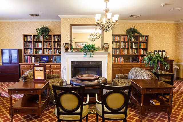 A cozy common room with chairs and tables arranged around a central fireplace, flanked by bookshelves and a large mirror.