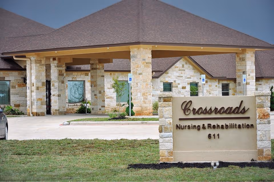 Exterior view of Crossroads Nursing and Rehabilitation facility showing a stone building with a covered entrance and a sign in front displaying the facility name and address number 611.