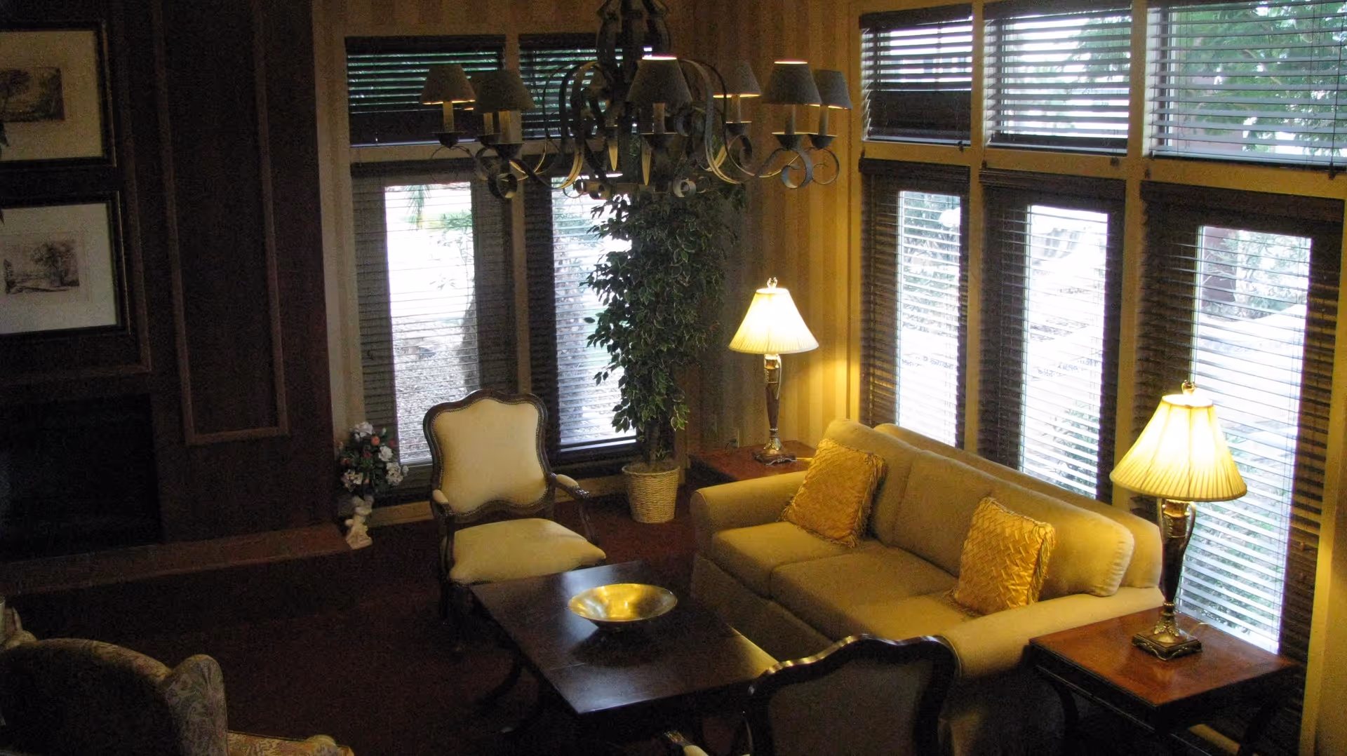 A cozy living room with a beige sofa adorned with two yellow pillows, two armchairs, a wooden coffee table with a decorative bowl, two table lamps with pleated shades on side tables, a large chandelier hanging from the ceiling, and large windows with blinds letting in natural light. There is also a potted plant near the windows and framed artwork on the wall.