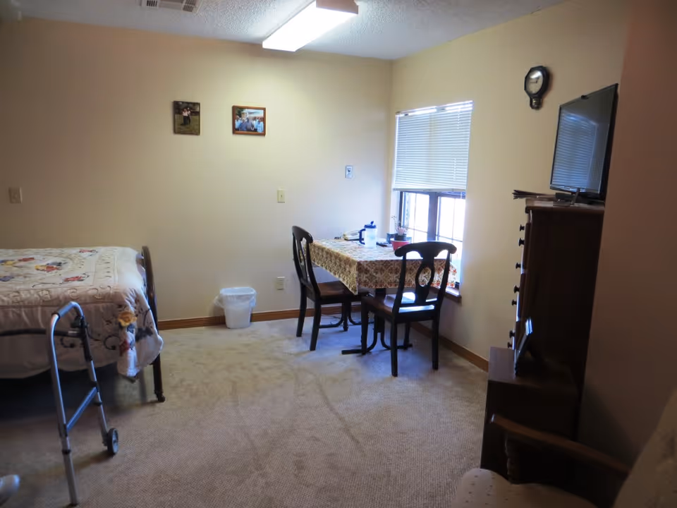 A small assisted living room with a single bed covered with a floral quilt, a walker beside the bed, a small table with two chairs near a window with blinds, a wooden dresser with a TV on top, and a wall clock above the TV. The room has beige walls and carpeted floor.