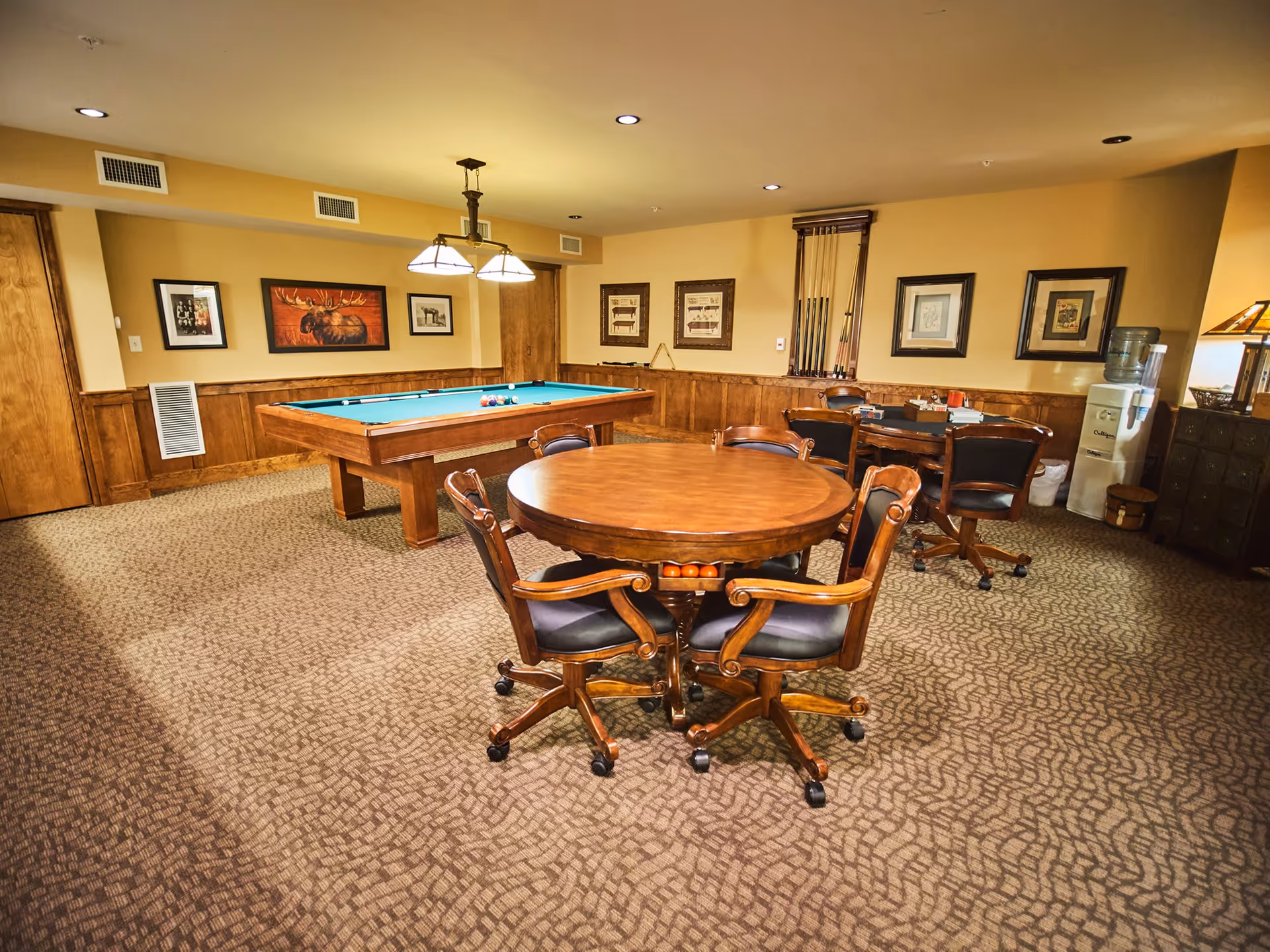 A cozy game room with a pool table and two round wooden tables surrounded by chairs on casters. The walls are decorated with framed pictures and a rack holding pool cues. There is a water cooler and a lamp on a cabinet in the corner. The room has warm lighting and carpeted flooring.