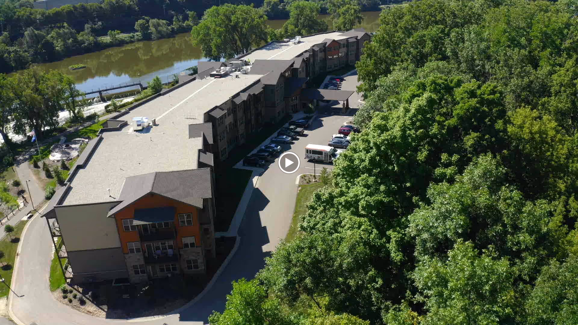 Aerial view of a multi-story senior living building by a river, surrounded by trees and a parking lot.