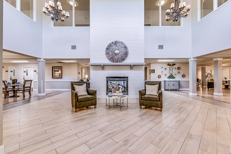 Spacious senior living facility common area with a large white brick fireplace in the center, flanked by two cushioned wicker chairs and a small round table with decorative items. The room features light wood flooring, high ceilings with two chandeliers, and visible adjacent dining and seating areas.