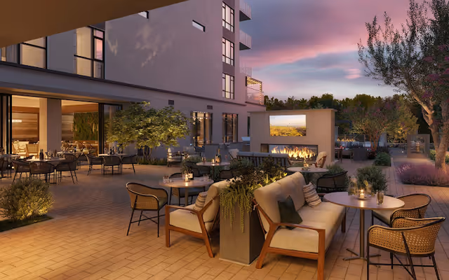 Outdoor patio area at dusk with comfortable seating including cushioned chairs and sofas around small tables. There is a modern outdoor fireplace with a built-in screen, surrounded by plants and trees. The patio is adjacent to a building with large windows showing an indoor dining area.