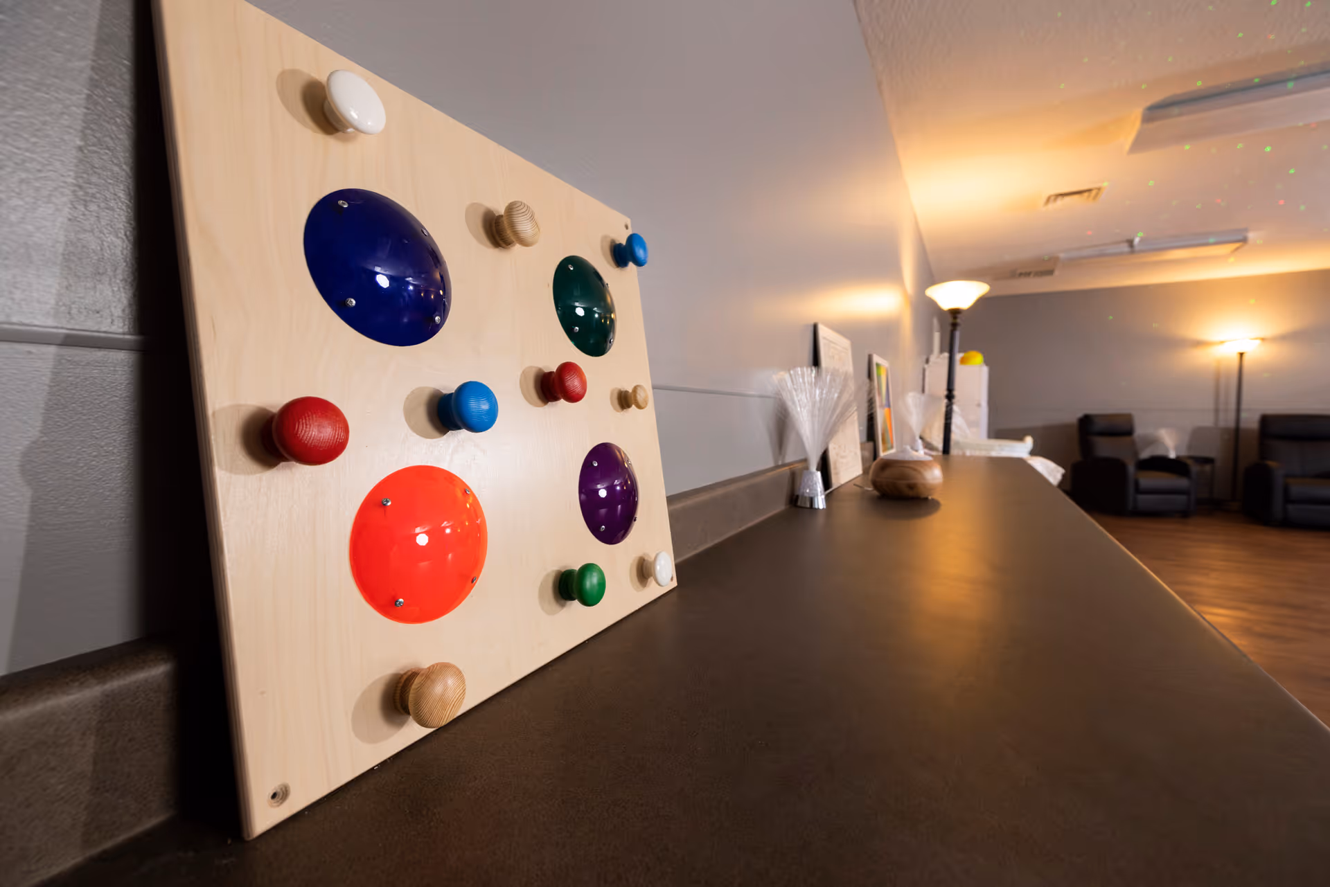 A sensory activity board with various colorful knobs and shapes mounted on a wooden panel, placed on a countertop in a softly lit room with comfortable chairs and lamps in the background.