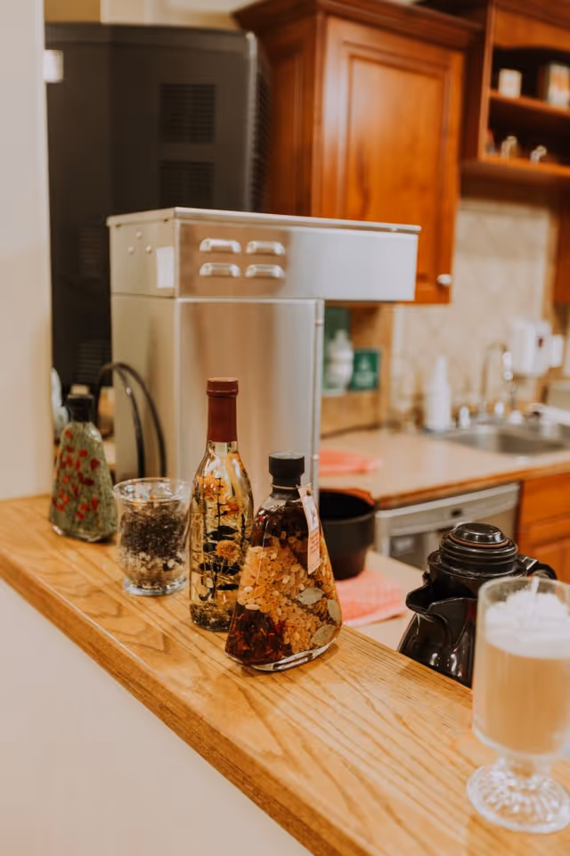 A kitchen counter with decorative bottles filled with herbs and spices, a glass container with coffee beans, a coffee maker, and a glass of frothy coffee. In the background, there are wooden cabinets, a sink, and a dishwasher.