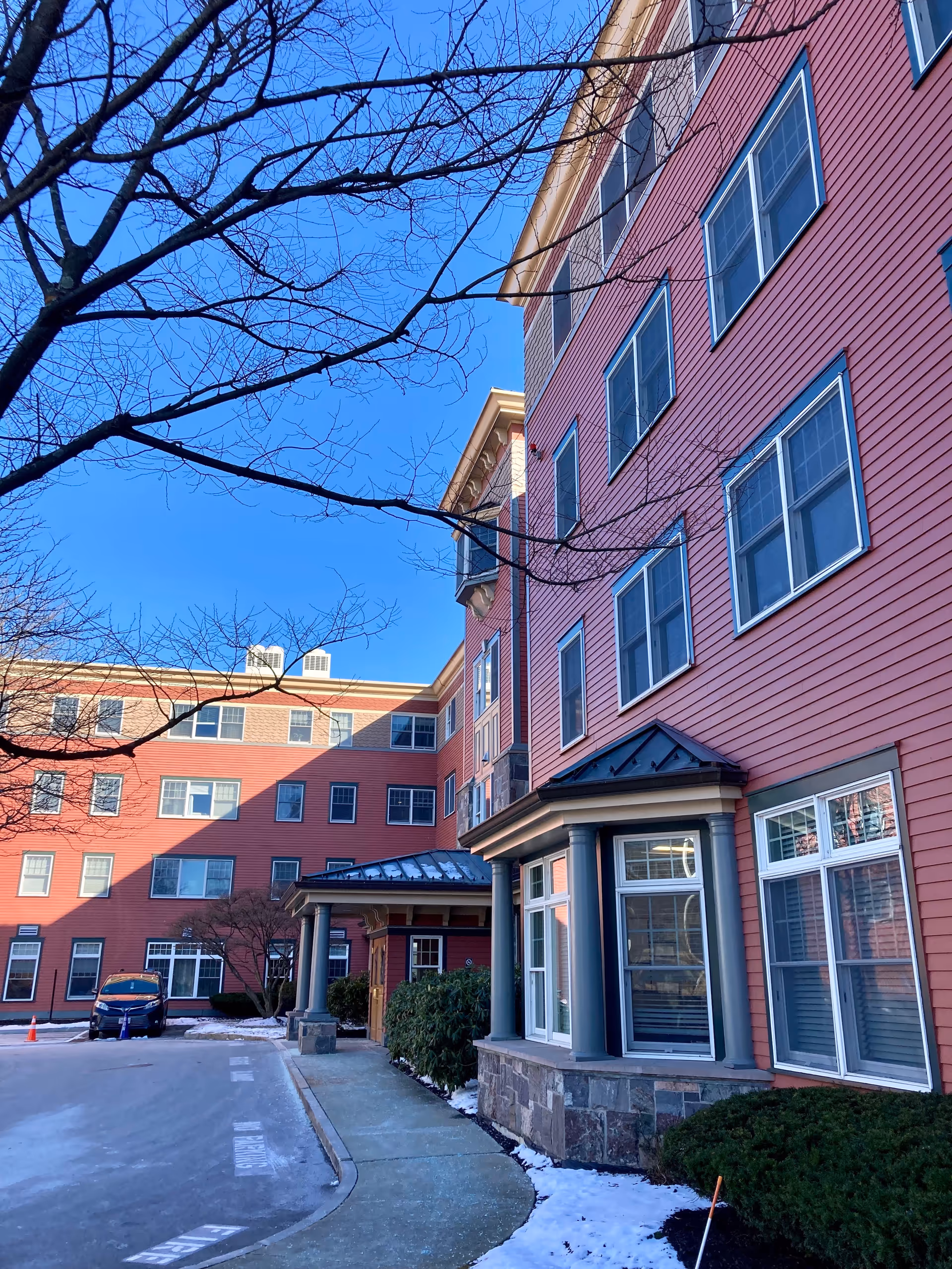 Exterior view of a multi-story senior living facility building with red siding and multiple windows. There is a sidewalk and some bushes in front, with patches of snow on the ground and a clear blue sky above. A bare tree branch extends across the top of the image.