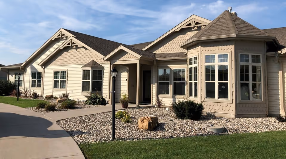 Front exterior of a beige single-story residential building with bay windows, a paved walkway, lamp posts, and rock landscaping.