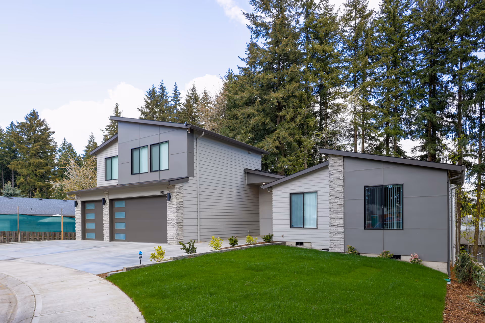 Modern gray two-story residential building with attached garage, driveway, and a green lawn in front with tall trees behind.