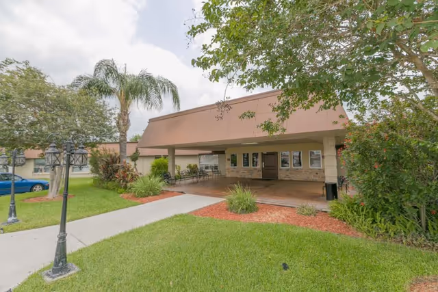 Covered entrance and drop-off area of a rehabilitation facility with a lawn, palm trees, and a sidewalk.