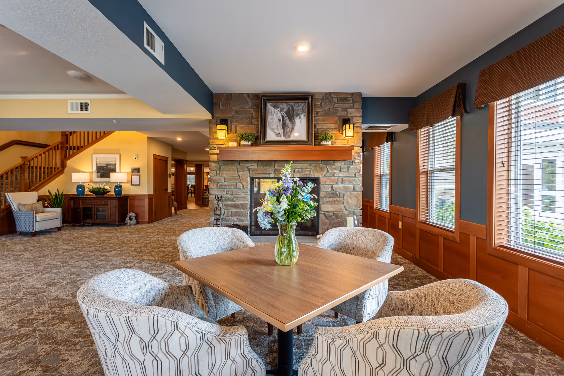 A cozy senior living common area with a square wooden table surrounded by four upholstered chairs. A vase with a colorful flower arrangement sits on the table. Behind the table is a stone fireplace with a framed black and white horse picture above it, flanked by two wall-mounted lantern-style lights. To the right, large windows with wooden blinds and brown valances let in natural light. To the left, a staircase and a wooden sideboard with lamps and plants are visible in the background.