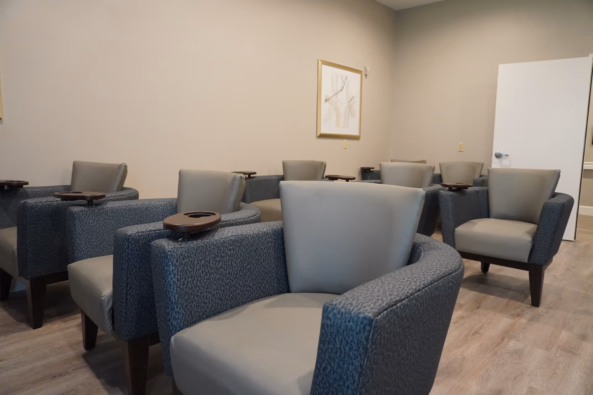A room with multiple gray upholstered armchairs arranged in rows, each chair featuring a small attached wooden tray. The room has light-colored walls, a framed abstract artwork, and a door slightly ajar in the background. The floor is covered with light wood-style flooring.
