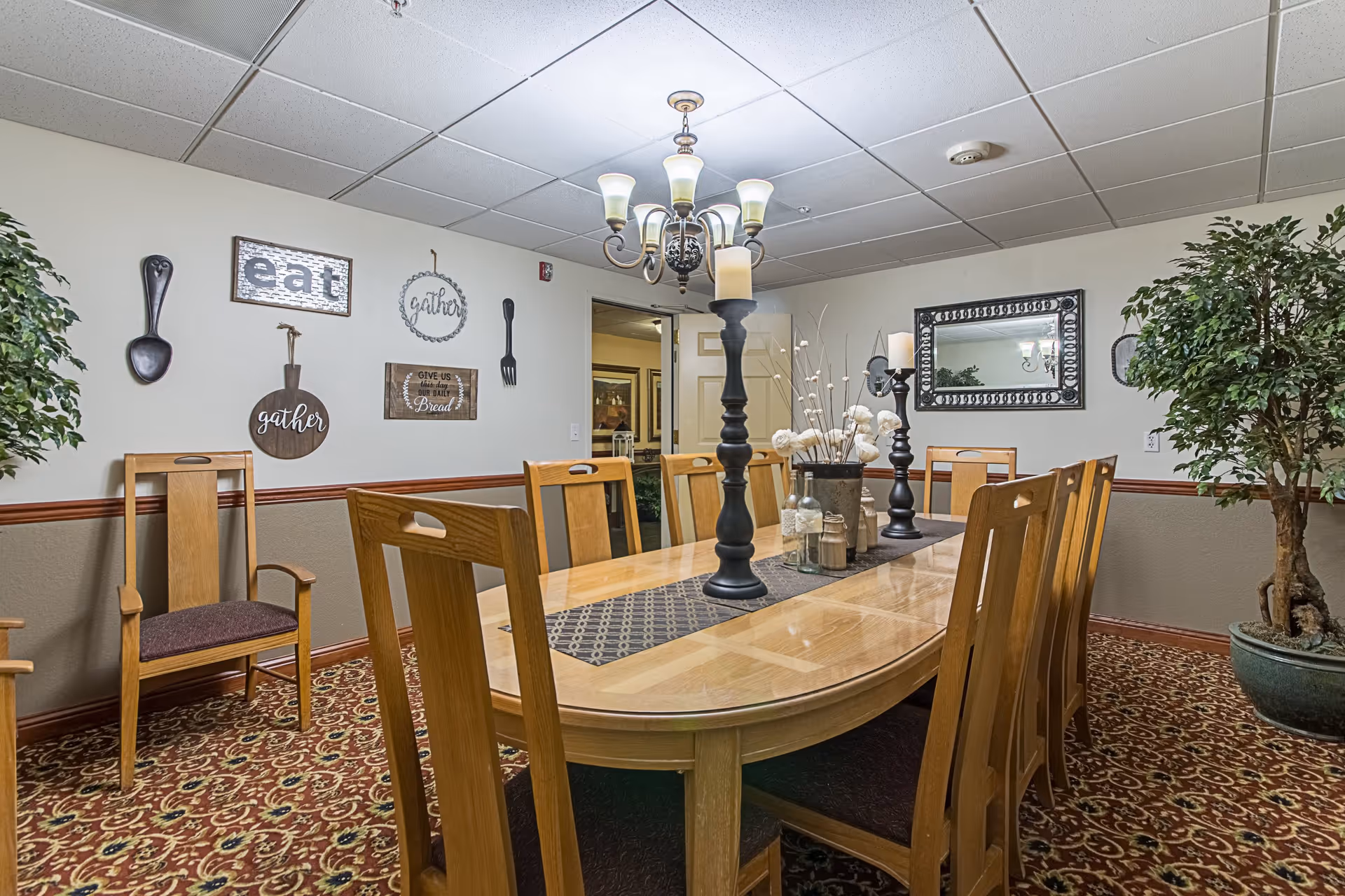 A dining room with a long wooden table surrounded by wooden chairs with dark cushions. The table is decorated with a patterned runner, two black candle holders with candles, and a vase with white flowers. The room has patterned carpet flooring, beige walls with a brown chair rail, and a ceiling with white tiles. Wall decorations include large wooden utensils, framed signs with words like 'eat' and 'gather', and a large mirror. There are two large potted plants in the corners of the room.
