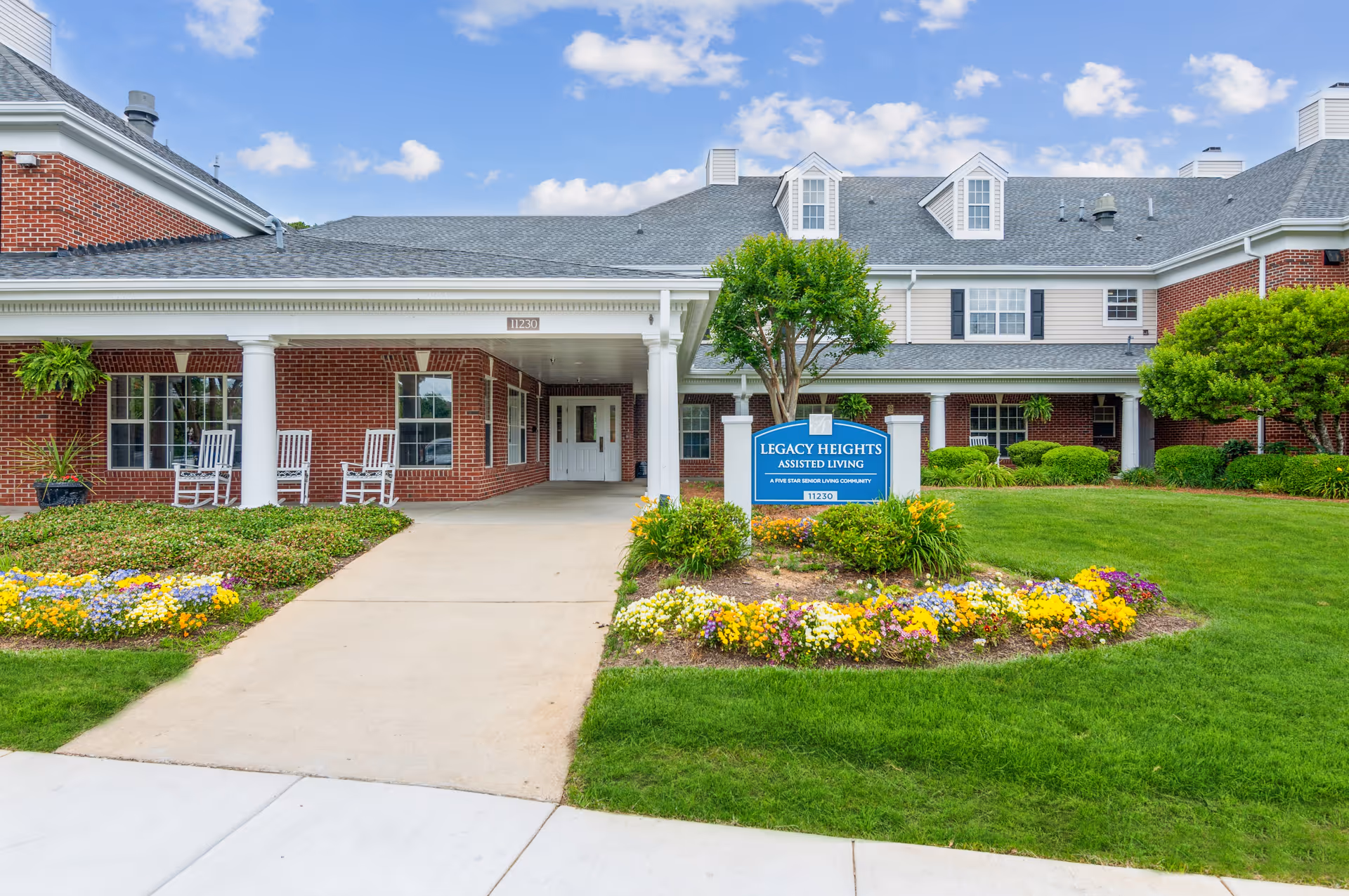 Front exterior view of Legacy Heights Senior Living Community building with a covered entrance, white columns, red brick walls, white rocking chairs on the porch, well-maintained green lawn, colorful flower beds, and a blue sign displaying the facility name.