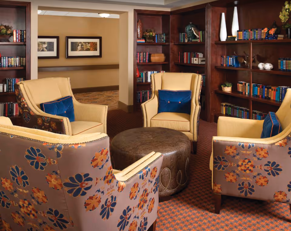 A cozy seating area with four upholstered armchairs arranged around a round leather ottoman. The chairs have a floral pattern on the back and yellow cushions with blue accent pillows. Behind the seating area are built-in wooden bookshelves filled with books and decorative items. The room has warm lighting and a carpeted floor with a patterned design.