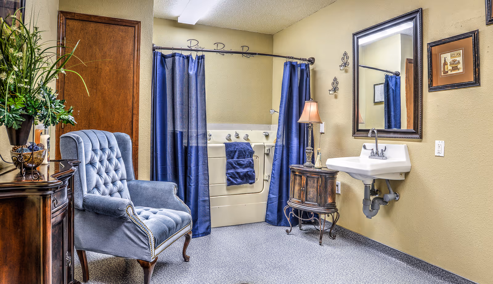 A decorated accessible bathroom showing a walk-in bathtub with navy curtains, a wall-mounted sink and mirror, and a tufted armchair with a side table.