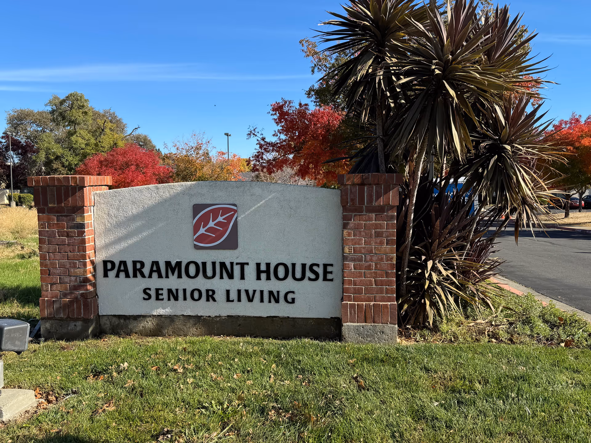 Outdoor view of a sign for Paramount House Senior Living, featuring a white stone panel with black text and a red leaf logo, flanked by two brick pillars. Surrounding the sign are green grass, palm-like plants, and trees with autumn-colored leaves under a clear blue sky.