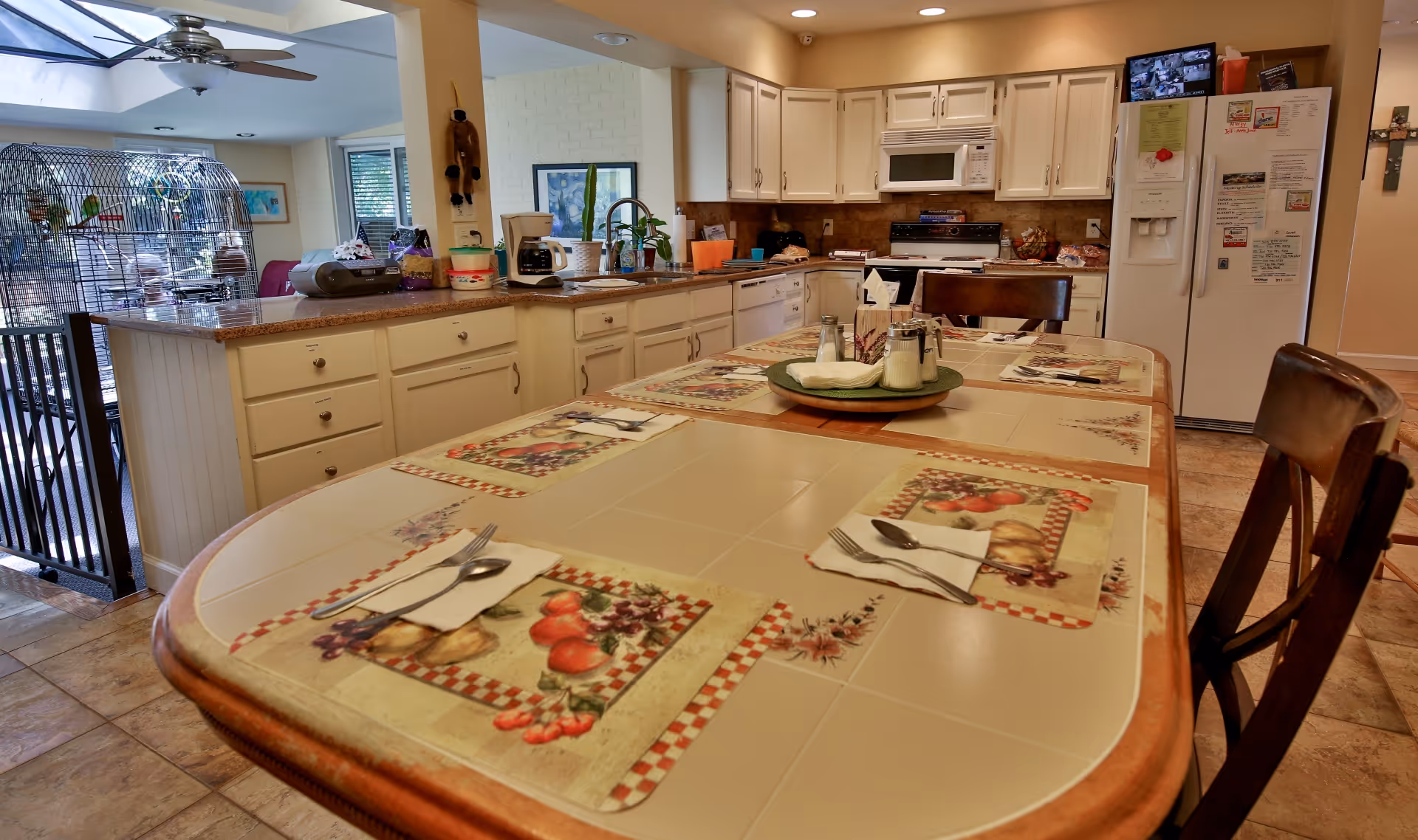 A kitchen and dining area with a tiled dining table set with placemats, napkins, and utensils. The kitchen has white cabinets, a microwave, stove, dishwasher, and a refrigerator with notes and magnets. A birdcage with birds is visible on the left side near a window, and a ceiling fan is above.
