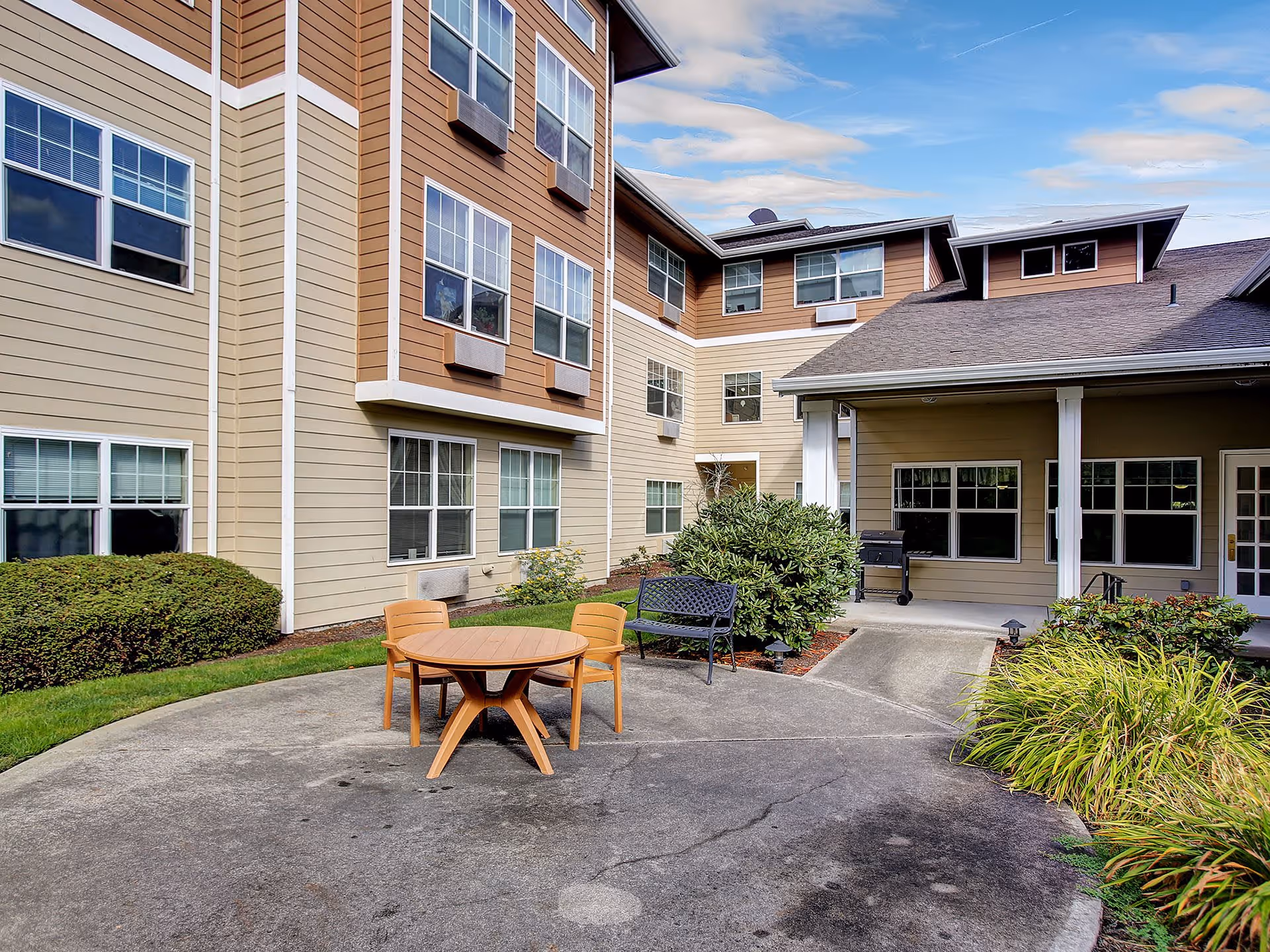 Outdoor patio area at Prestige Senior Living Rosemont featuring a round wooden table with two chairs, a black metal bench, shrubs, and a covered porch attached to a multi-story building with beige and brown siding and multiple windows.