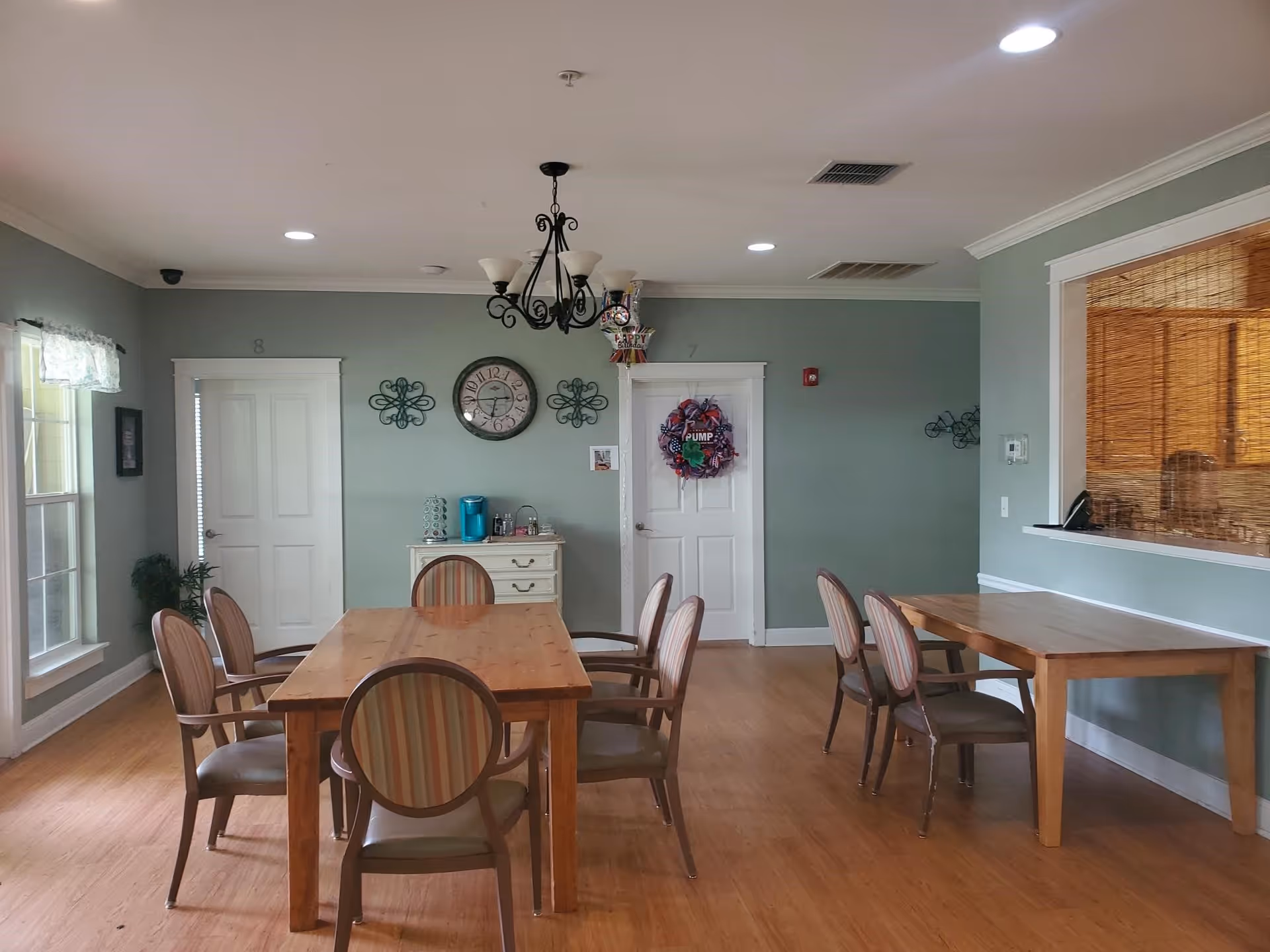 A dining room with two wooden tables and several chairs with striped upholstery. The walls are painted light green with white trim. There is a clock and decorative wall hangings on the far wall, along with two white doors, one decorated with a wreath. A chandelier hangs from the ceiling, and there is a window with a bamboo shade on the right side. The floor is wooden.