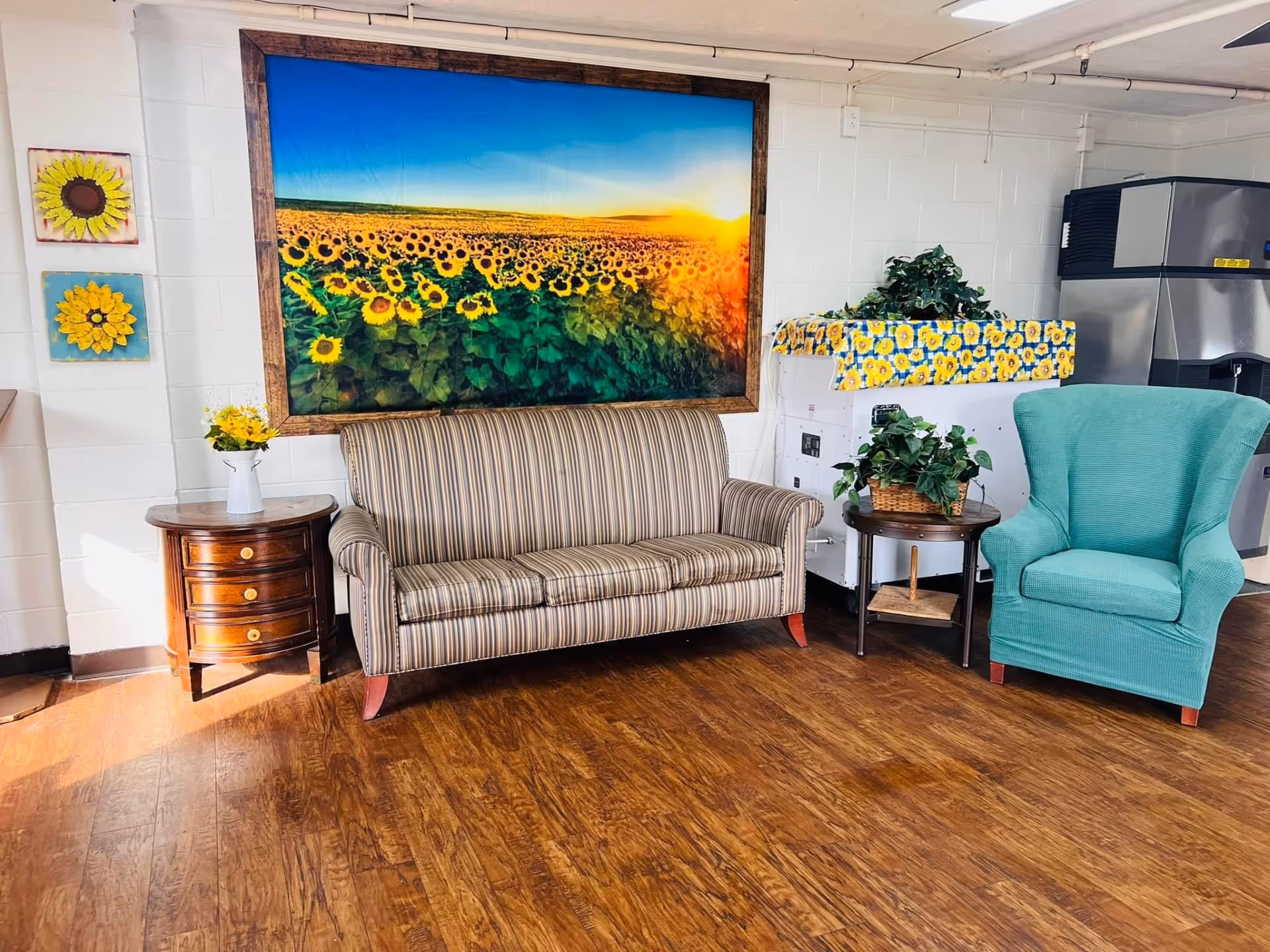 A seating area with a striped sofa, teal armchair, side tables, plants, and a large sunflower landscape artwork on the wall.
