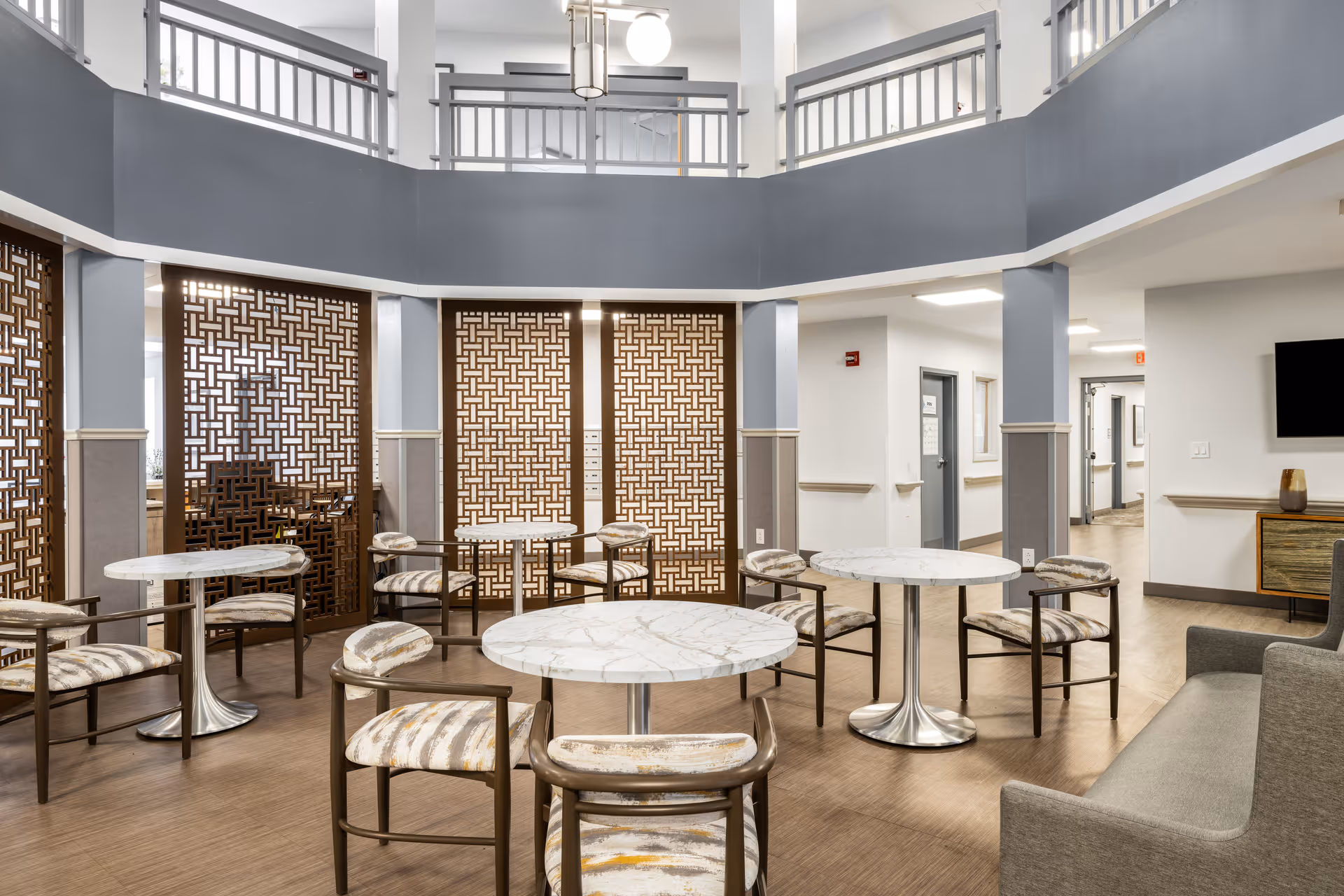 A spacious senior living community common area with round marble-top tables and cushioned chairs arranged around the room. The space features decorative wooden lattice panels, a gray and white color scheme, and a two-story ceiling with a balcony railing above. There is a gray sofa on the right side and a hallway leading to other rooms in the background.