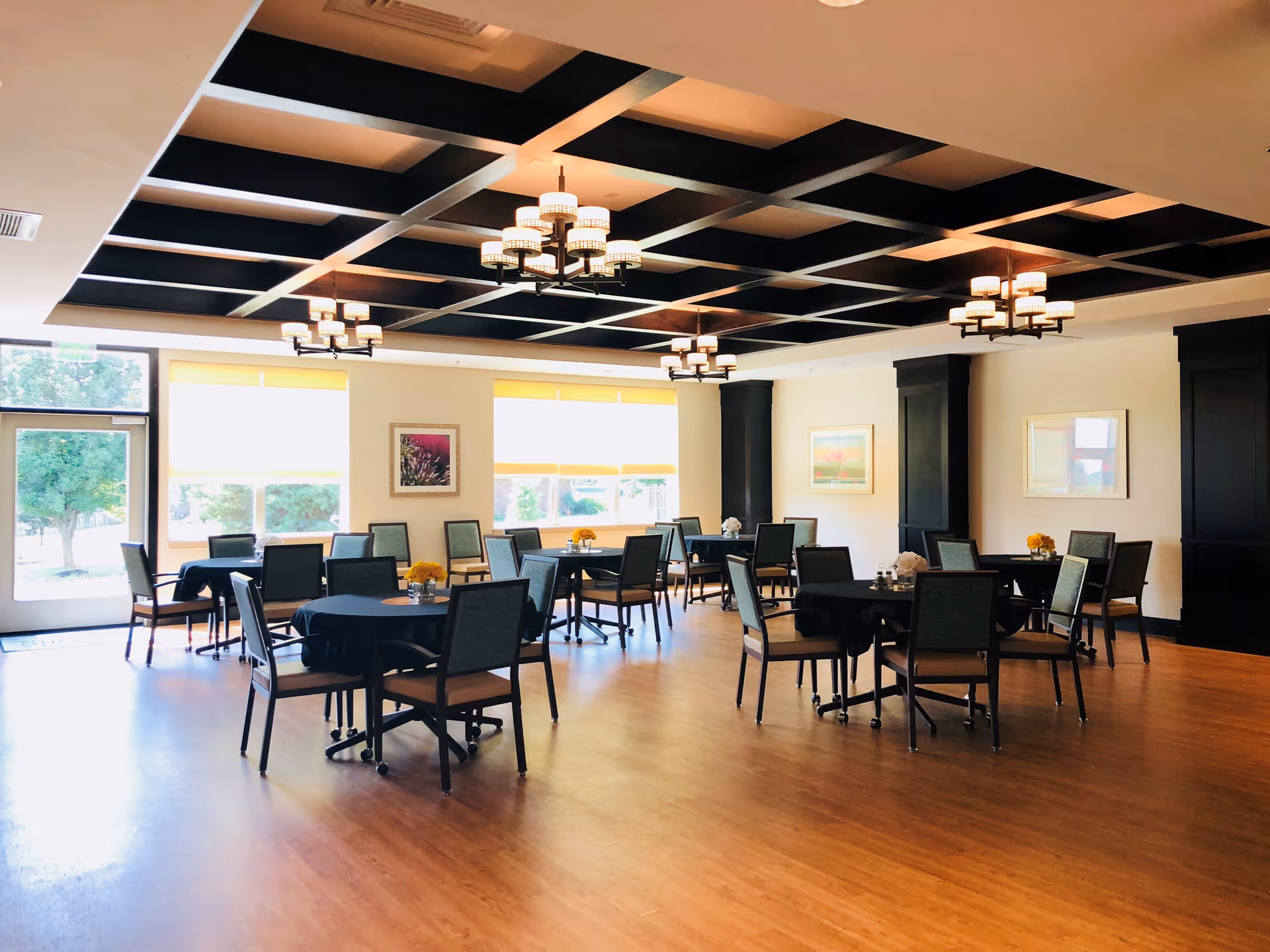 Spacious dining room with round tables and chairs, large windows, and a decorative coffered ceiling with chandeliers.