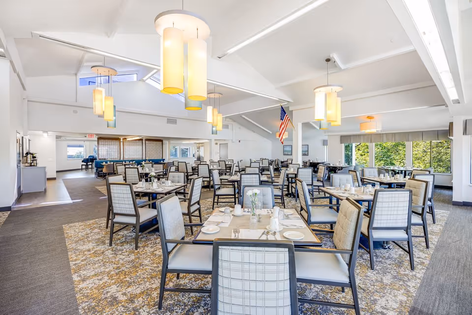 A spacious and bright dining room with multiple tables and chairs neatly arranged. The tables are set with plates, cups, and silverware, and small flower vases are placed on each table. Large windows on the right side allow natural light to fill the room, and several modern pendant lights hang from the ceiling. An American flag is displayed near the center of the room.