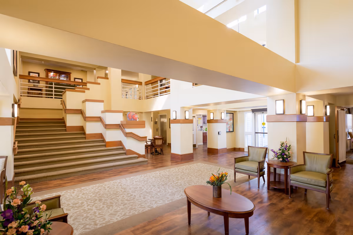 A spacious and well-lit interior common area with wooden flooring, beige walls, and a patterned carpet. There are two green armchairs and a wooden coffee table with a flower arrangement in the foreground. A staircase with carpeted steps and wooden railings leads to an upper level. The area is decorated with wall-mounted lights and additional seating and tables are visible in the background.