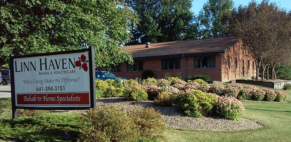 Exterior view of a single-story brick building surrounded by landscaped bushes and trees under a clear blue sky. In the foreground, there is a large sign that reads 'Linn Haven Rehab & Healthcare, Where Caring Makes the Difference, 641-394-3151, Rehab to Home Specialists.'