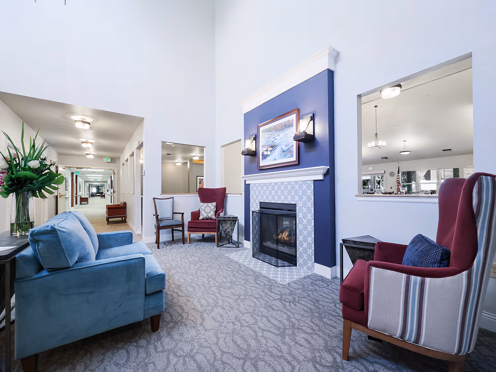 A cozy seating area in a senior living facility featuring a blue sofa, two armchairs with one in burgundy and the other striped, a fireplace with a blue accent wall and a framed picture above it, and a long hallway with additional seating and lighting fixtures.