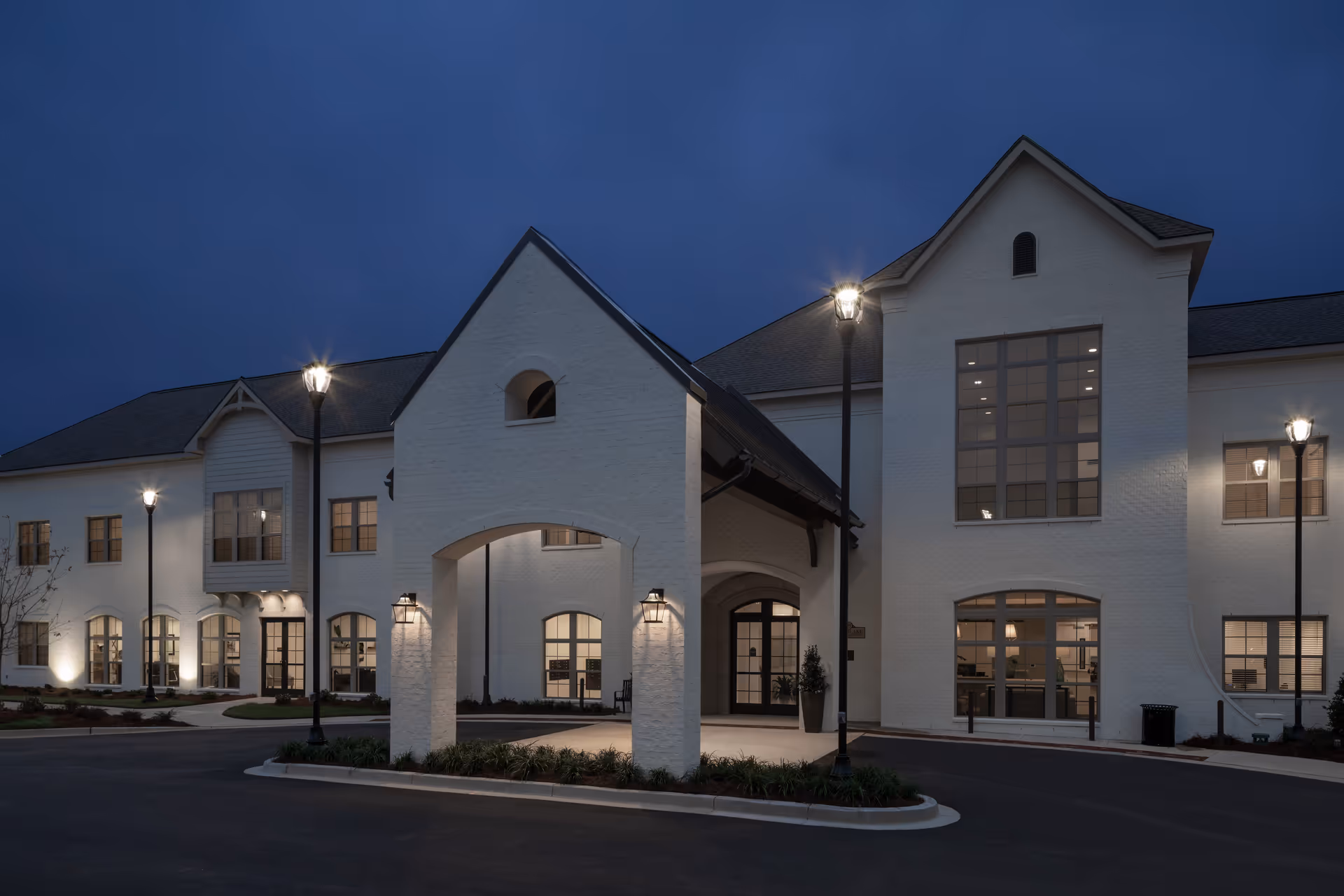 Exterior nighttime view of The Blake at Hollingsworth Park building with white brick walls, large windows, a covered entrance, and illuminated street lamps along the driveway.