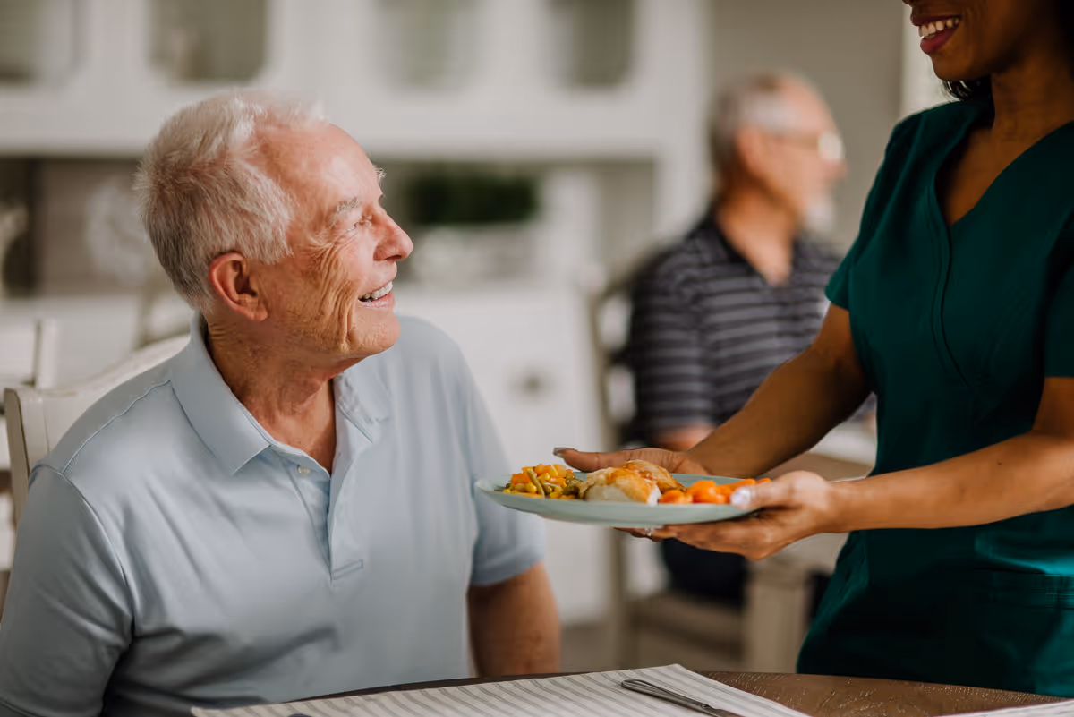A smiling elderly man seated at a dining table is being handed a plate of food by a caregiver.