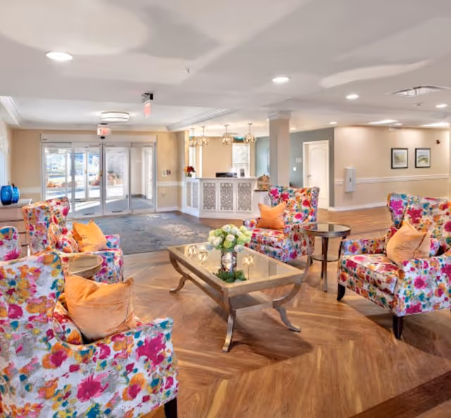 Bright assisted living lobby with floral armchairs arranged around a glass-top coffee table and a reception desk in the background.