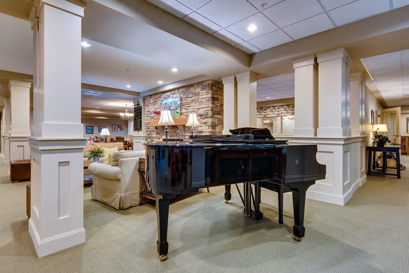 Interior view of a senior living facility lounge area featuring a black grand piano in the foreground, beige upholstered armchairs, a stone fireplace with a painting above it, and soft lighting from table lamps and ceiling lights.