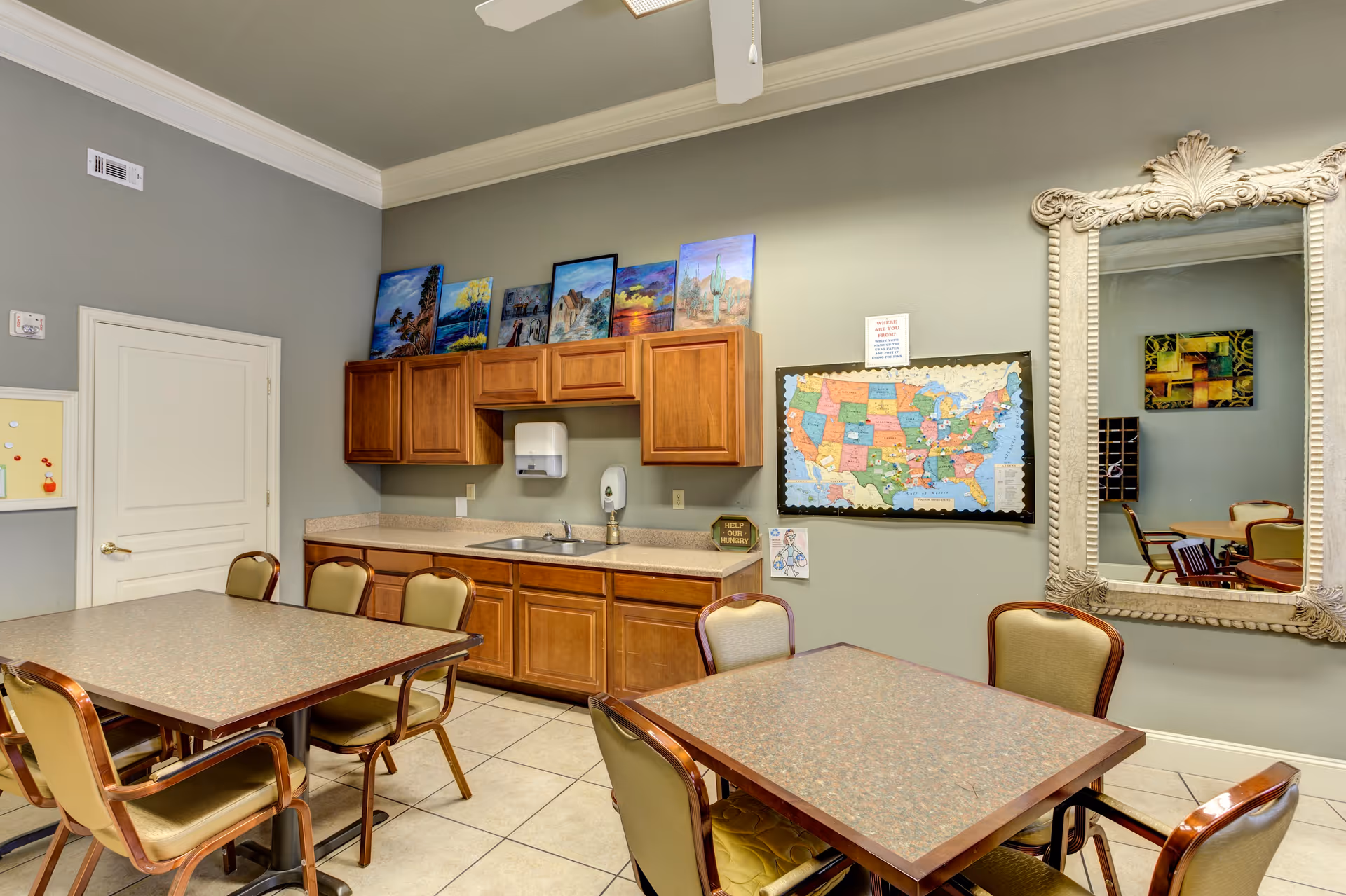 A common area with two tables surrounded by chairs, a countertop with a sink and wooden cabinets above and below. On the wall above the countertop are several colorful paintings and a large map of the United States. A large ornate mirror is mounted on the adjacent wall reflecting part of the room.