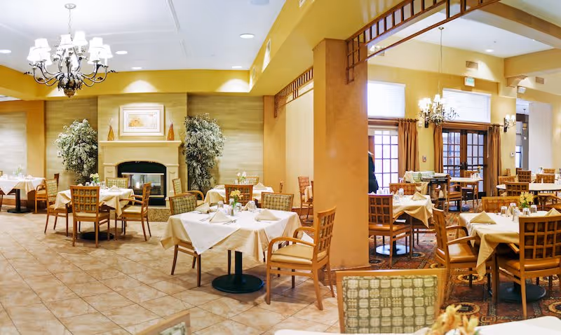 Dining room with neatly set tables and chairs, chandeliers, and a fireplace in a warm-toned interior.