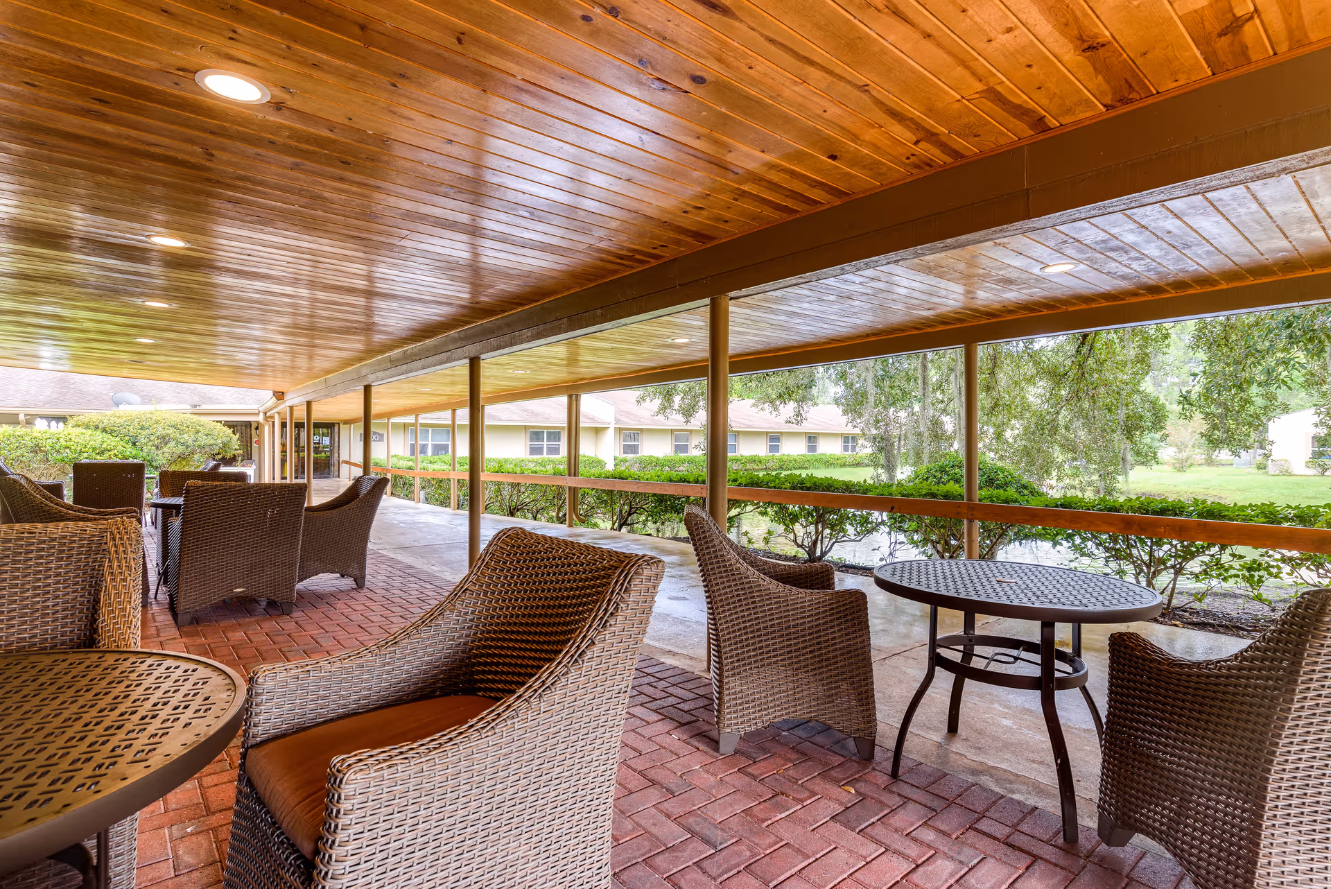 Covered outdoor patio area with wicker chairs and round metal tables on a brick floor, overlooking green bushes and trees with a building in the background.