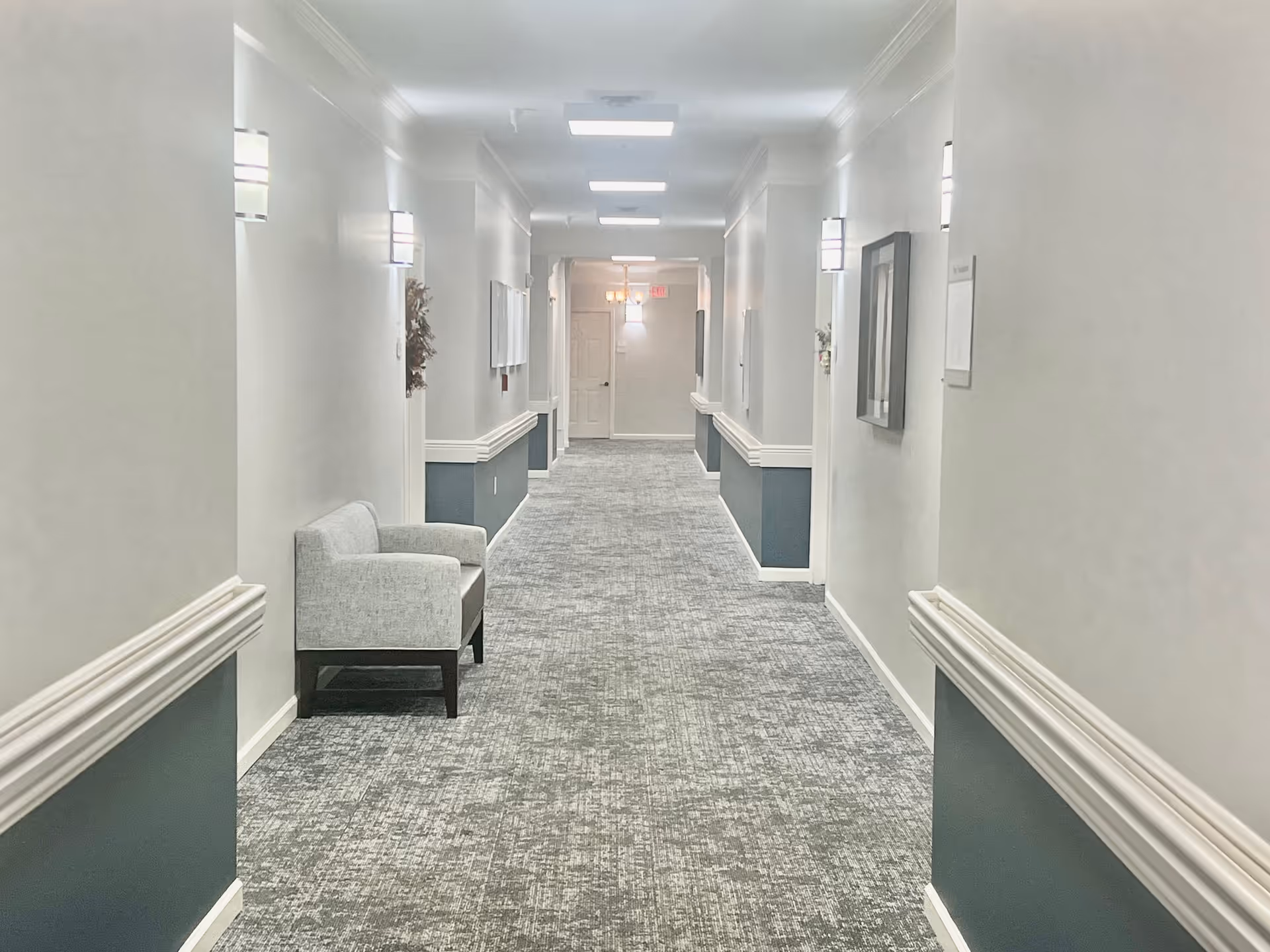 A long, well-lit hallway in a senior living facility with light gray walls and blue wainscoting. The carpet is patterned in shades of gray. There are wall-mounted lights evenly spaced along the corridor, a single gray upholstered chair on the left side, and framed artwork on the walls. A door is visible at the end of the hallway.