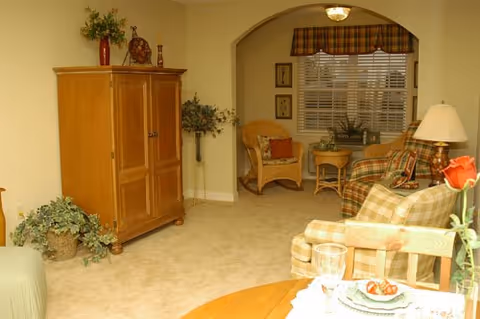 A cozy living room area with beige carpet, a wooden armoire, wicker chairs with cushions, a small round table, and a window with blinds and a plaid valance. There is a table with a lamp and a plaid sofa, and a dining table with a plate of food and a glass of water in the foreground.
