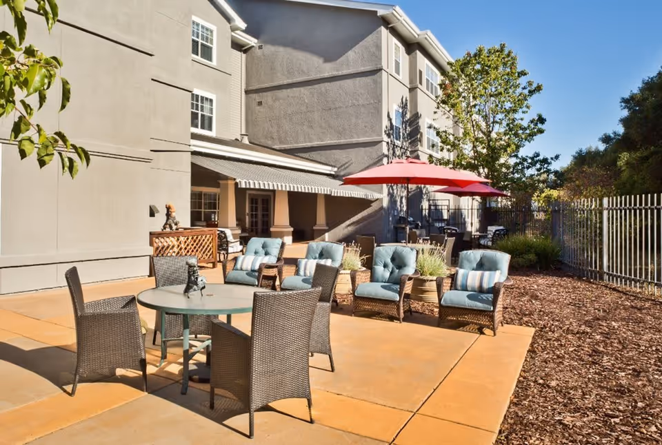 Outdoor patio area at a senior living facility with a round glass table surrounded by four wicker chairs, and a seating area with four cushioned armchairs and striped pillows. There is a large red umbrella providing shade, a building with multiple windows in the background, and some trees and plants along the fenced perimeter.