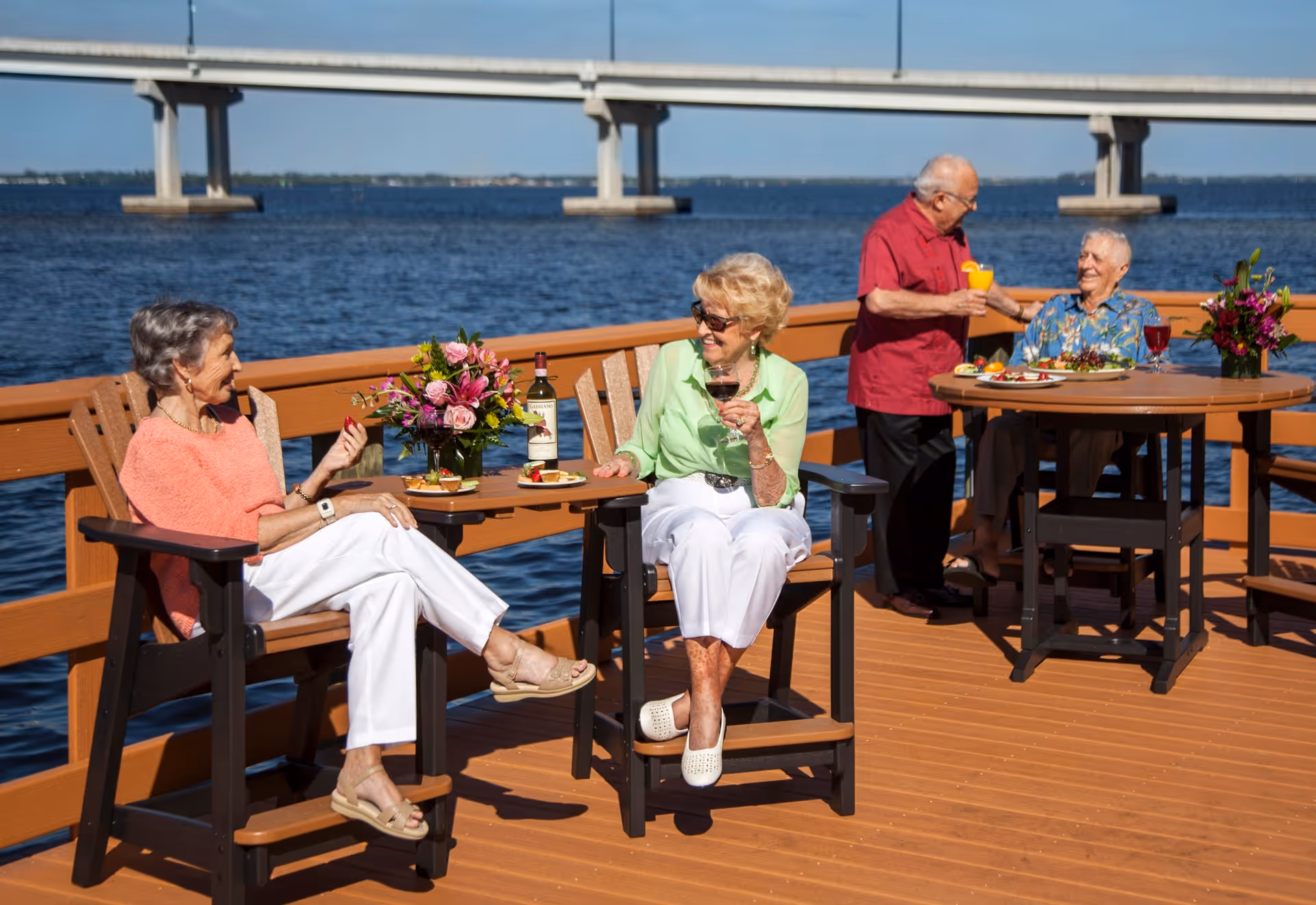 Four elderly people enjoying drinks and snacks on a wooden deck by the water with a bridge in the background. Two women are seated at a small table with flowers and wine, while two men are at a round table with drinks and food, engaging in conversation.