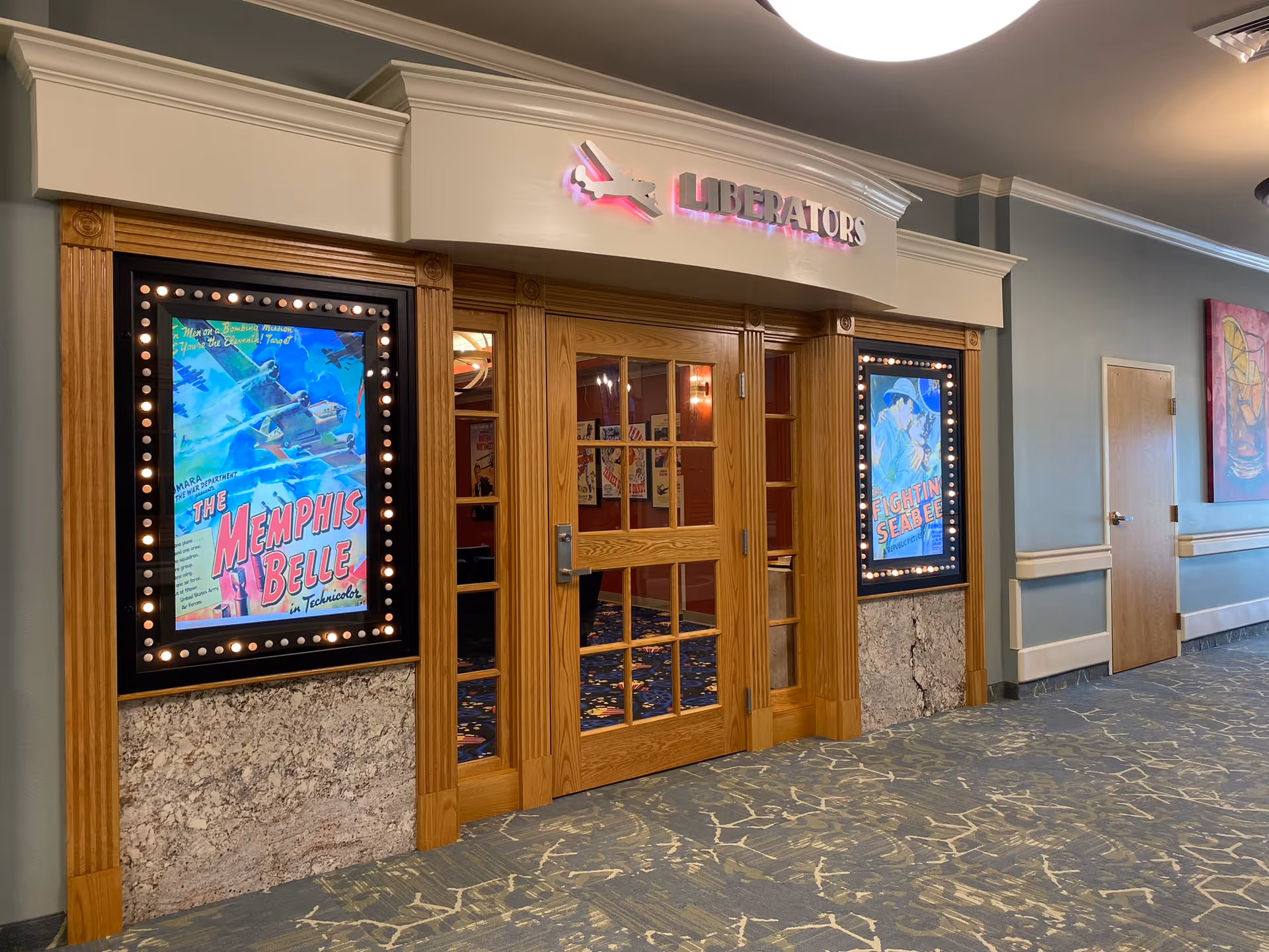 Entrance to a room named 'Liberators' with wooden double doors and movie poster-style lighted frames on either side. The posters display vintage war-themed movie titles 'The Memphis Belle' and 'Fightin Seabee'. The hallway has patterned carpet and light blue walls with a closed wooden door and a colorful abstract painting on the right.