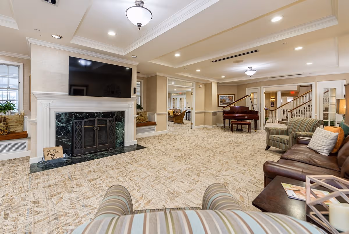 A spacious and well-lit living room area in a senior living facility featuring a large flat-screen TV mounted above a white fireplace with a dark green marble surround. The room has beige walls, patterned carpet, and comfortable seating including a leather sofa, striped armchairs, and built-in window benches with cushions. A grand piano is positioned near the back wall, and there are multiple doorways leading to other rooms. Ceiling lights and a central light fixture illuminate the space.
