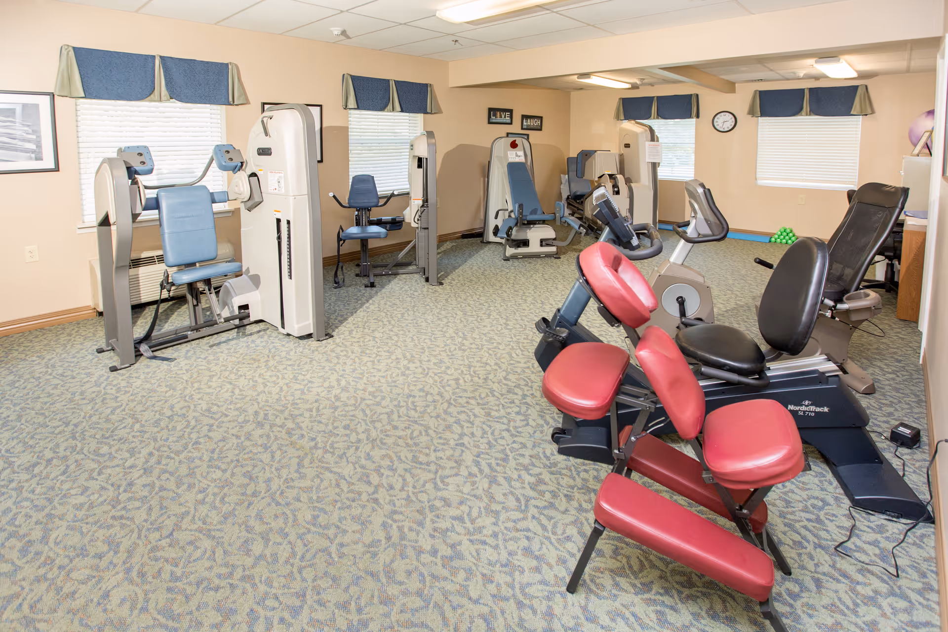 A fitness room in a senior living facility with various exercise machines including a red padded massage chair, stationary bikes, and strength training equipment. The room has beige walls, patterned carpet, windows with blue valances, and framed pictures on the walls.