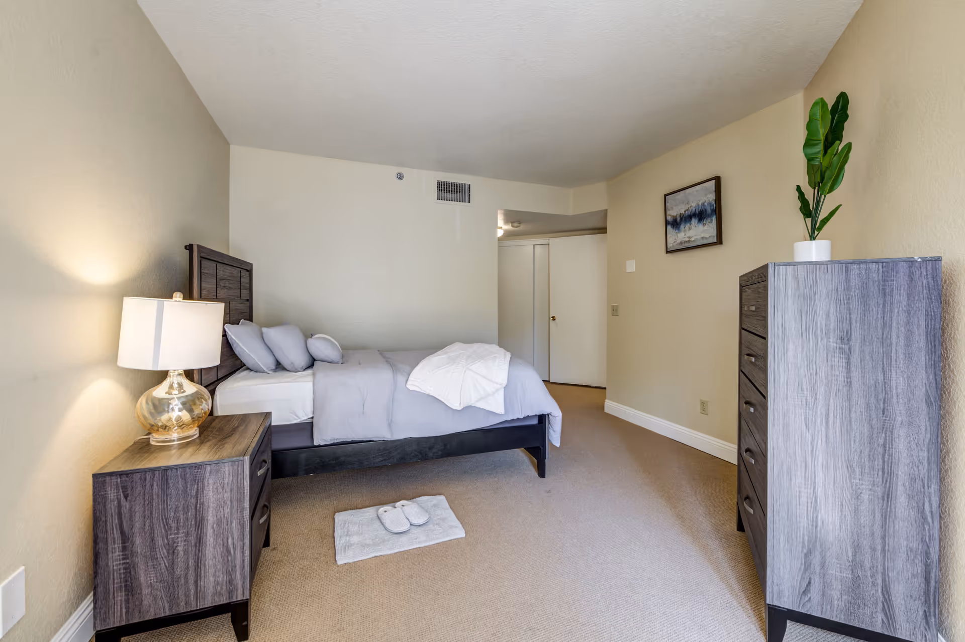 A neatly arranged bedroom with a bed made with gray bedding and pillows. A wooden nightstand with a lamp is beside the bed. On the floor near the bed is a small rug with a pair of slippers. Across from the bed is a tall wooden dresser with a potted plant on top. The walls are painted beige, and there is a framed abstract painting hanging above the dresser. The room has beige carpet and a white door in the background.